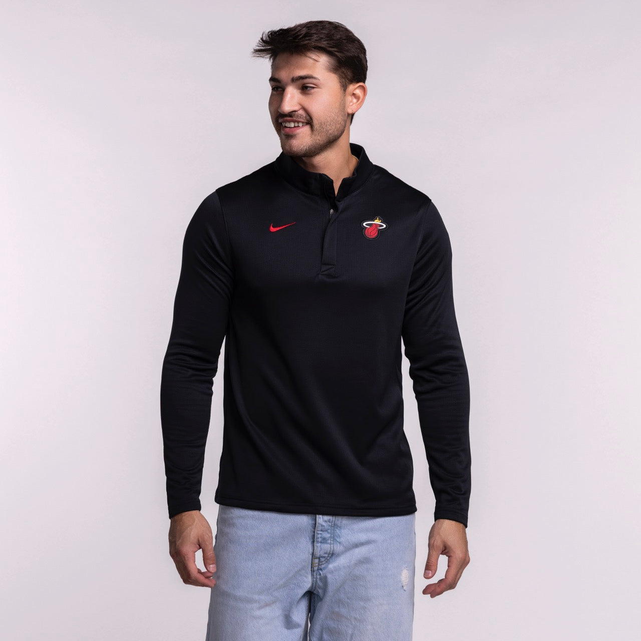 A man in light blue jeans smiles while wearing the Nike Miami HEAT Black Logo 1/4 Zip—a lightweight, sweat-wicking long-sleeve top with a red Nike logo and team emblem—standing against a plain light background.