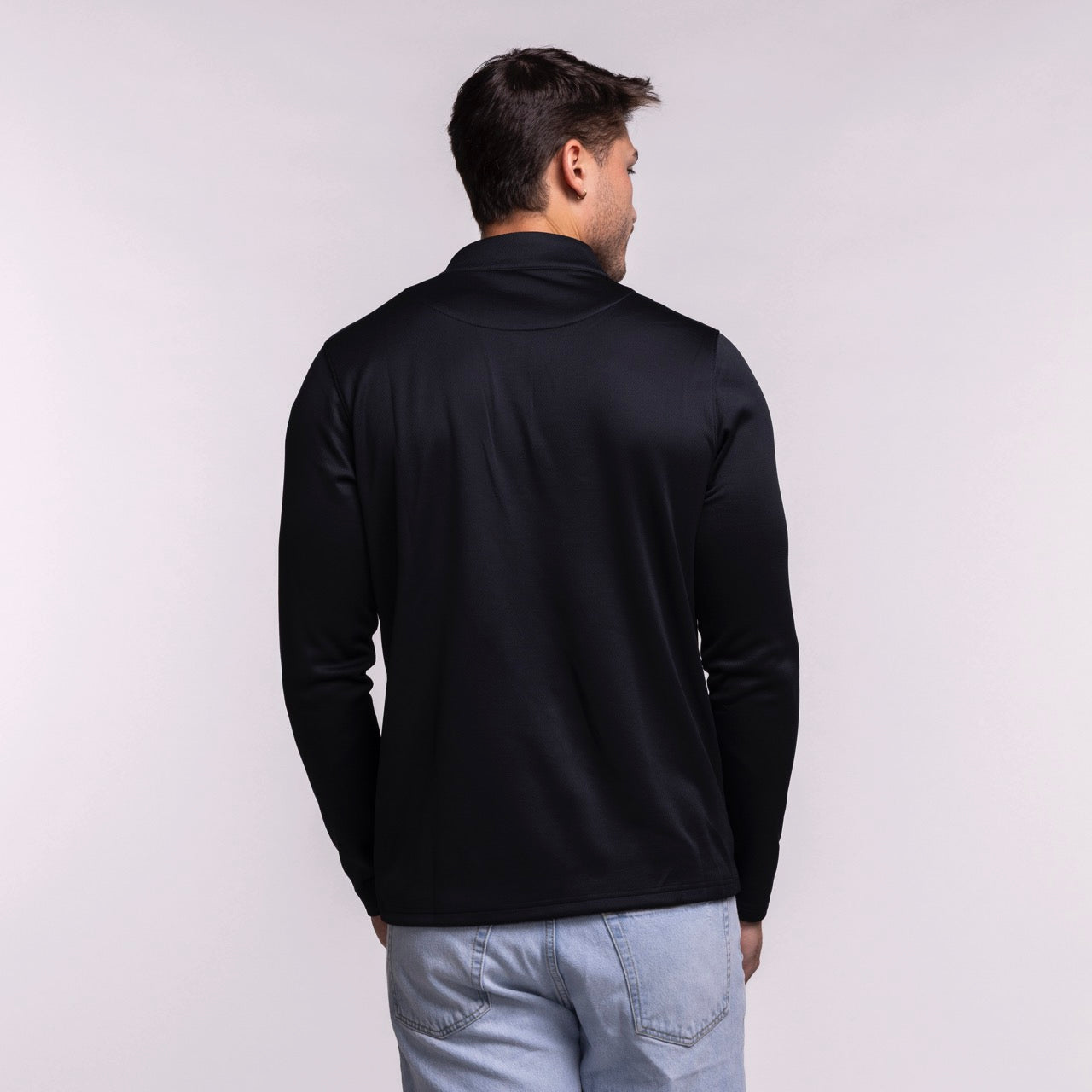 A man faces away from the camera, wearing a Nike Miami HEAT Black Logo 1/4 Zip top and light blue jeans against a plain, light-colored background.