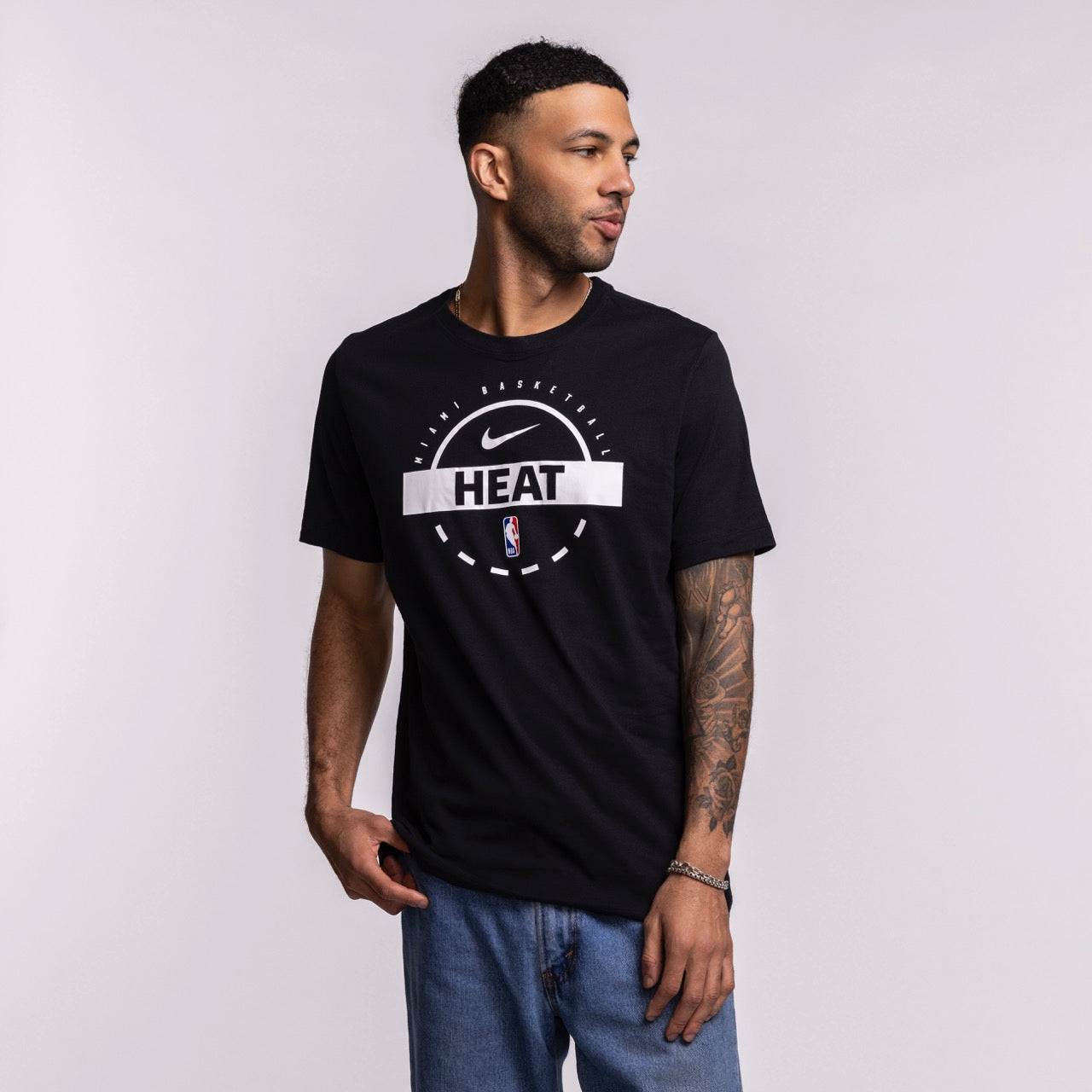 A man wearing the Nike Miami HEAT 2025-26 On-Court Practice Tee and blue jeans stands against a light background, looking right with one hand in his pocket and a tattoo sleeve visible on his left arm.