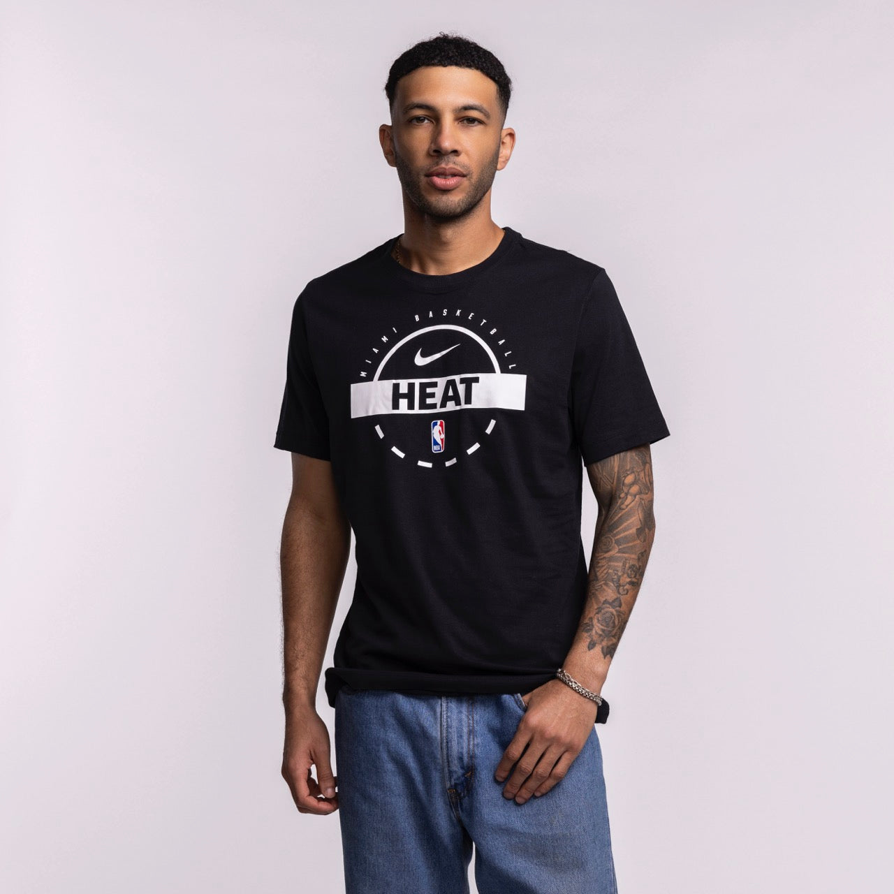 A man with short hair, a trimmed beard, and a tattoo sleeve on his left arm stands against a light background wearing blue jeans and the Nike Miami HEAT 2025-26 On-Court Practice Tee. One hand is in his pocket.