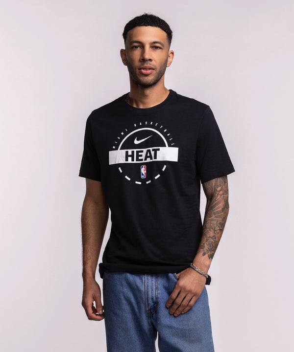 A man with short hair, a trimmed beard, and a tattoo sleeve on his left arm stands against a light background wearing blue jeans and the Nike Miami HEAT 2025-26 On-Court Practice Tee. One hand is in his pocket.