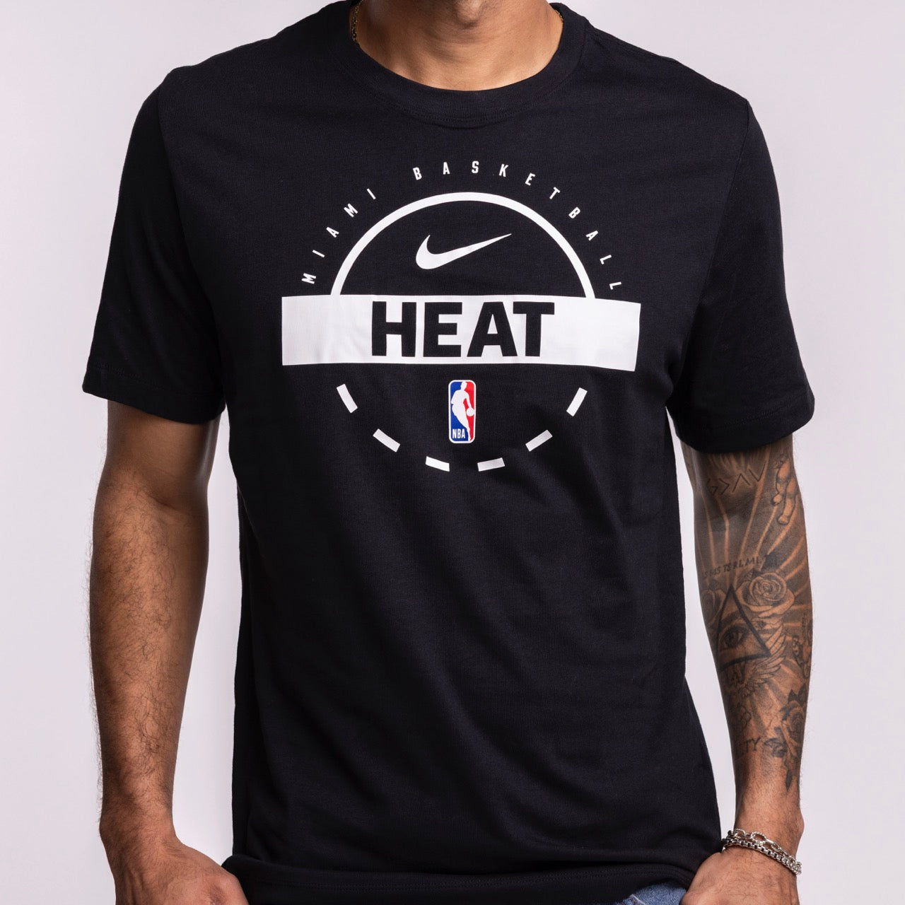 A person showcases a tattooed arm while wearing the Nike Miami HEAT 2025-26 On-Court Practice Tee by Nike, featuring the NBA logo on the front, against a plain background.