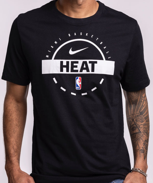 A man with short hair, a trimmed beard, and a tattoo sleeve on his left arm stands against a light background wearing blue jeans and the Nike Miami HEAT 2025-26 On-Court Practice Tee. One hand is in his pocket.