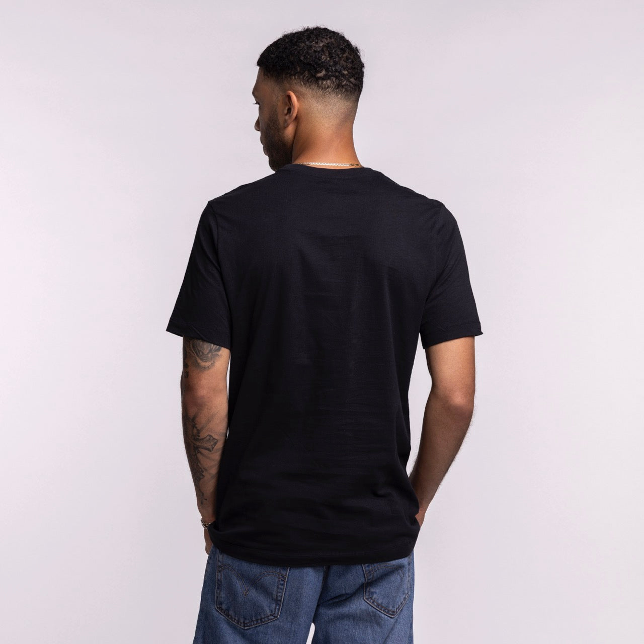 A man with short curly hair and arm tattoos faces away, wearing a Nike Miami HEAT 2025-26 On-Court Practice Tee by Nike and blue jeans, set against a light gray background.