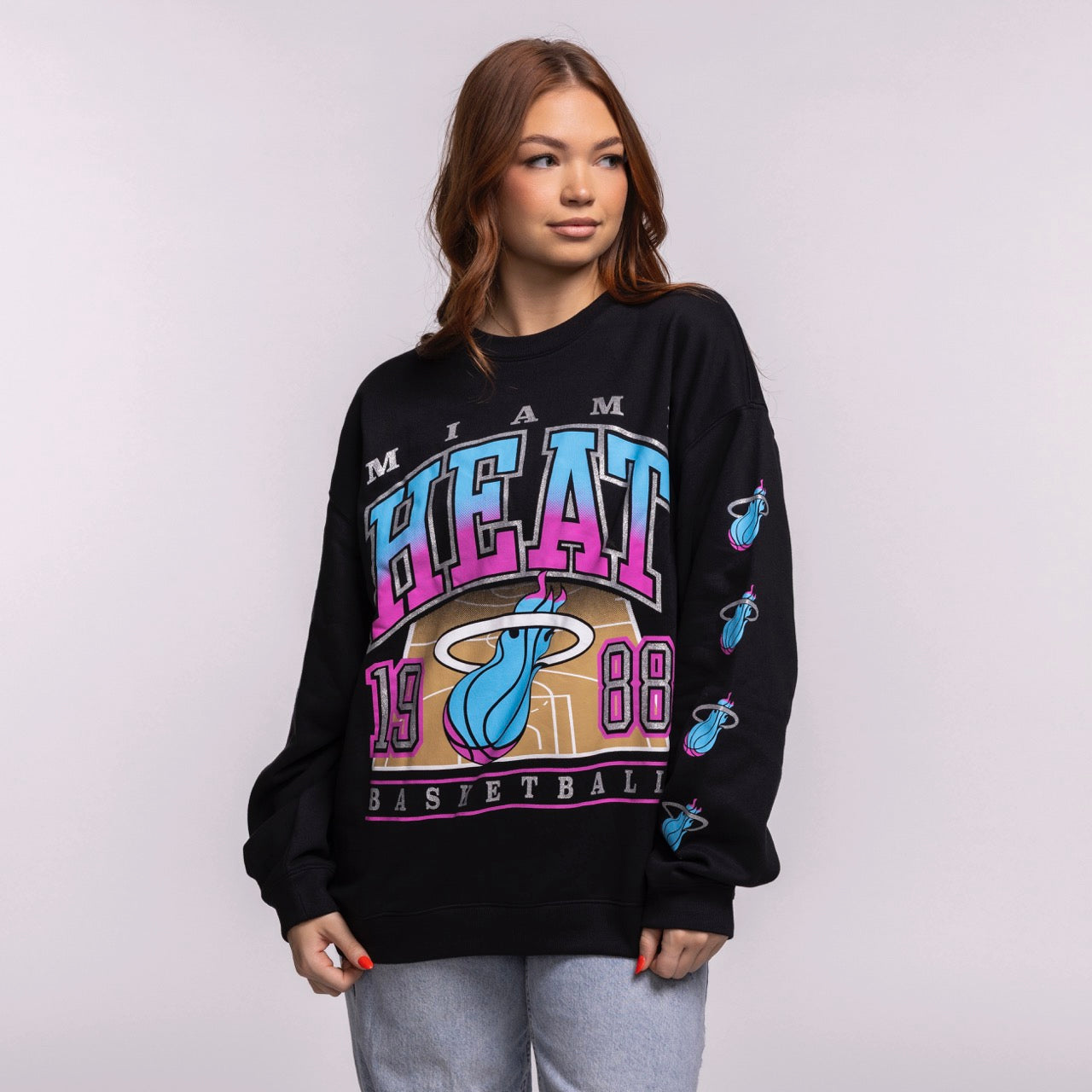 A young woman with long light brown hair wears a '47 Brand Miami HEAT Original Vice Nights 1988 Women's Crewneck, an oversized black sweatshirt with bold colorful graphics, paired with light blue jeans against a plain light background.
