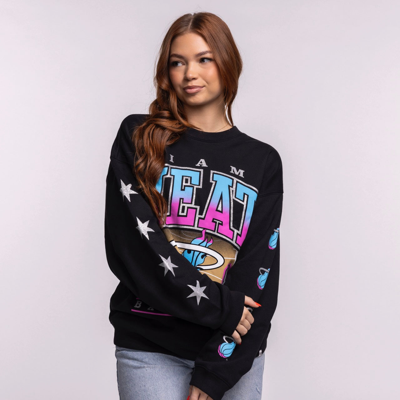 A young woman with long auburn hair models the '47 Brand Miami HEAT Original Vice Nights 1988 Women's Crewneck, featuring colorful graphics and white stars on the sleeve, against a plain light gray background.