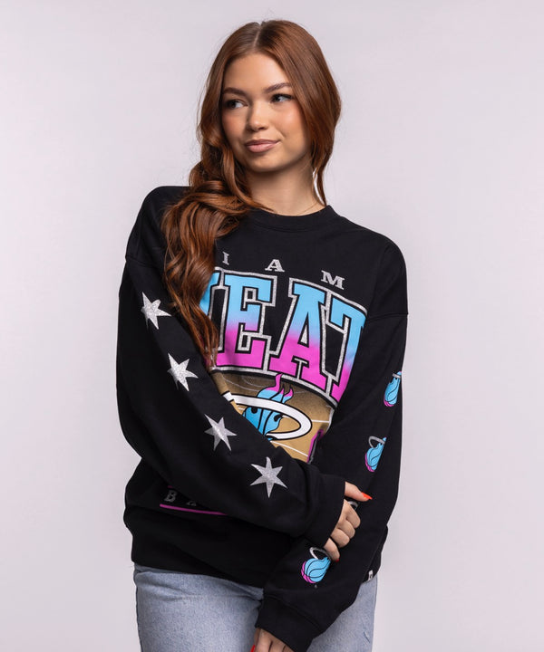 A young woman with long auburn hair models the '47 Brand Miami HEAT Original Vice Nights 1988 Women's Crewneck, featuring colorful graphics and white stars on the sleeve, against a plain light gray background.