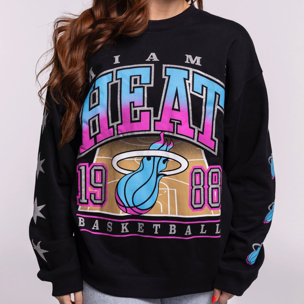 A person (head not visible) wears the '47 Brand Miami HEAT Original Vice Nights 1988 Women's Crewneck, an oversized black cotton-polyester sweatshirt featuring colorful HEAT text, a flaming basketball logo, 1988, and stars on the sleeves.