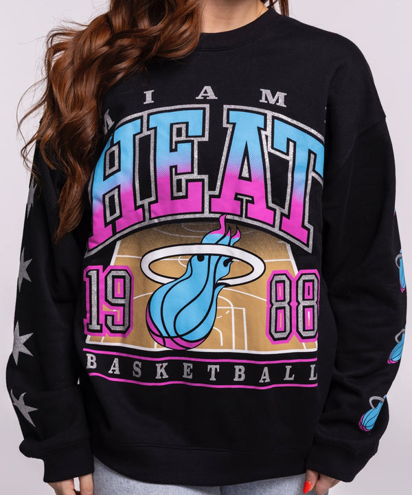 A young woman with long auburn hair models the '47 Brand Miami HEAT Original Vice Nights 1988 Women's Crewneck, featuring colorful graphics and white stars on the sleeve, against a plain light gray background.