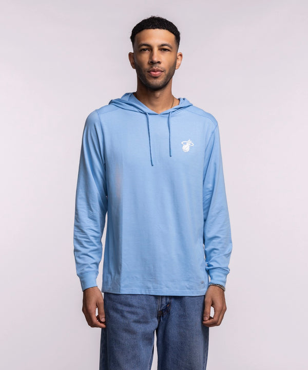 A man faces forward, wearing the '47 Brand Miami HEAT Original Vice Nights Blue Hoodie with a small white chest logo and blue jeans. The background is plain and light-colored.