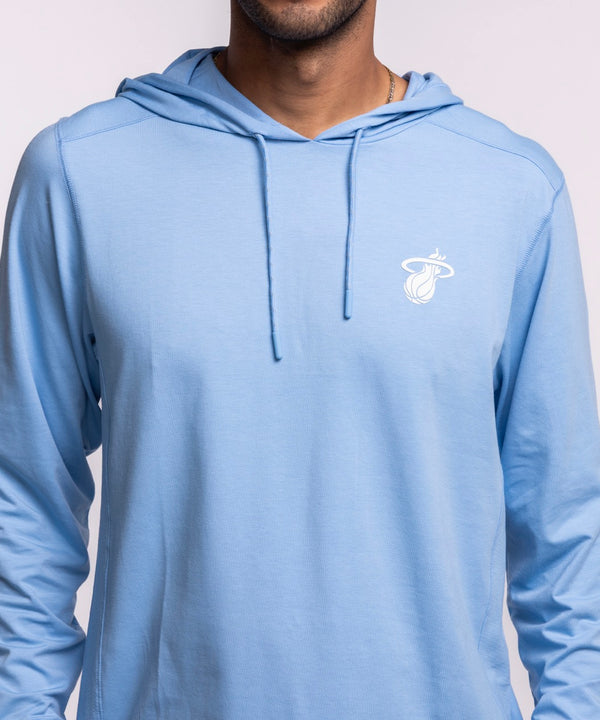 A man faces forward, wearing the '47 Brand Miami HEAT Original Vice Nights Blue Hoodie with a small white chest logo and blue jeans. The background is plain and light-colored.