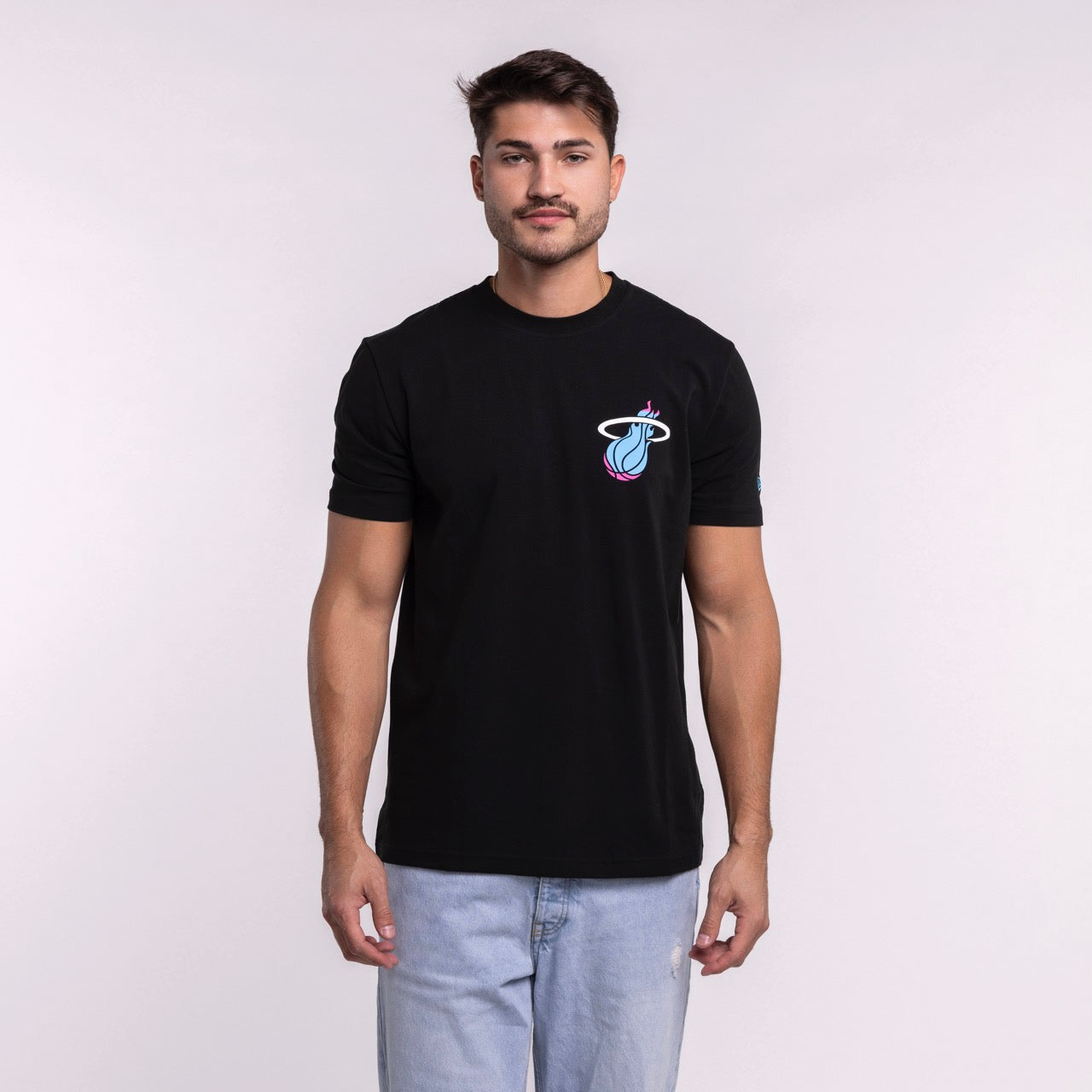 A man stands facing forward in a standard fit New Era Miami HEAT Original Vice Nights Back Print Tee with a colorful basketball logo on the chest and light blue jeans, showcasing effortless men's fashion against a plain light background.