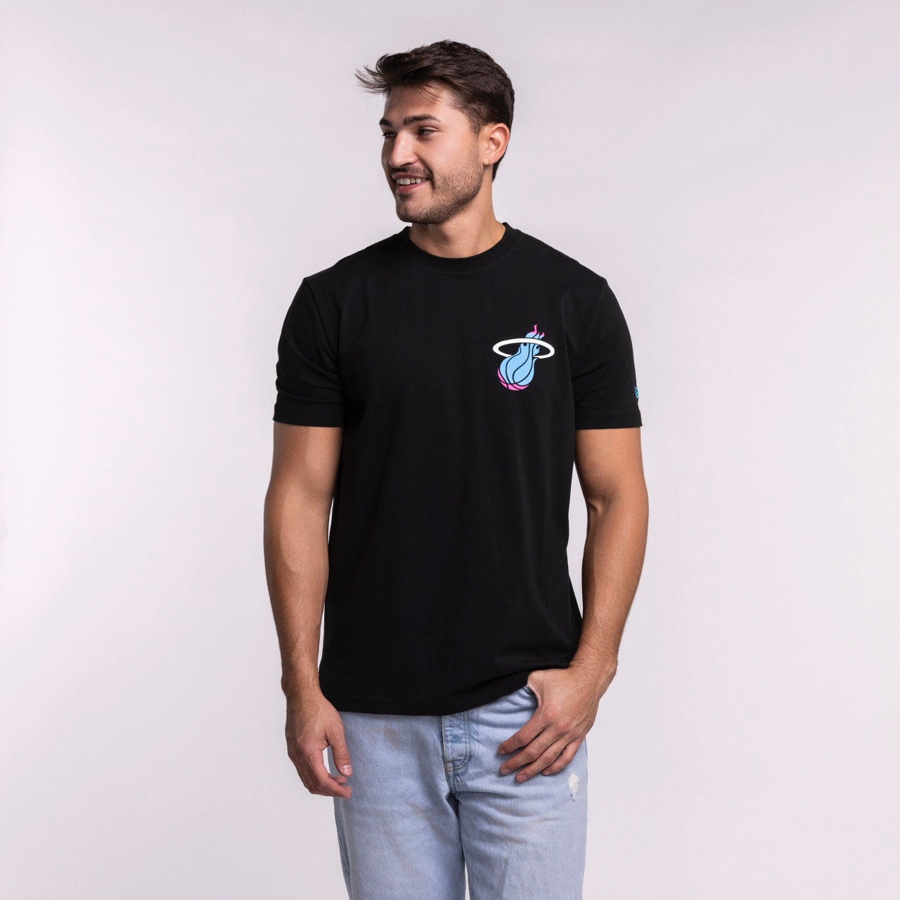 A man models the New Era Miami HEAT Original Vice Nights Back Print Tee, featuring a colorful basketball logo. He pairs it with light blue jeans and stands against a plain light background, smiling and looking to his left.