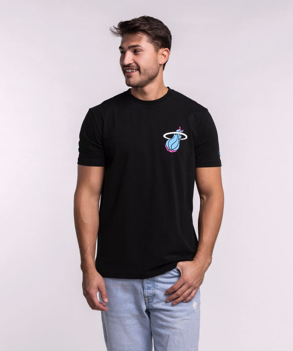 A man models the New Era Miami HEAT Original Vice Nights Back Print Tee, featuring a colorful basketball logo. He pairs it with light blue jeans and stands against a plain light background, smiling and looking to his left.