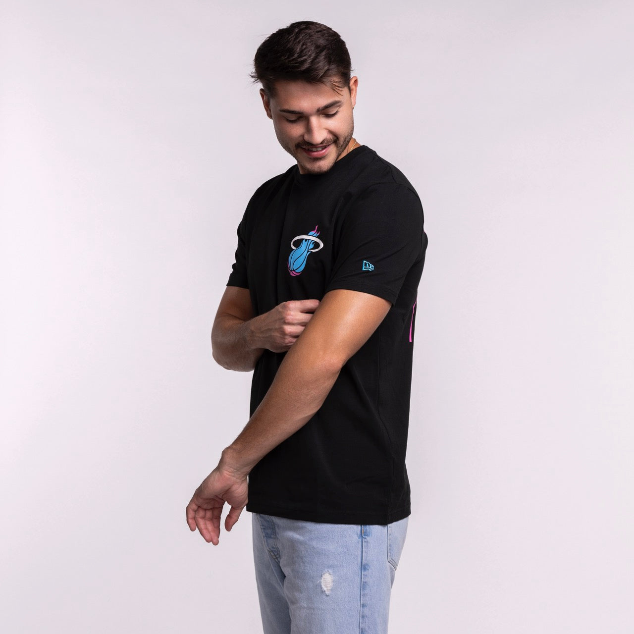 A man wearing the New Era Miami HEAT Original Vice Nights Back Print Tee in standard fit and light blue jeans adjusts his sleeve against a plain background, effortlessly highlighting stylish men's fashion by New Era.