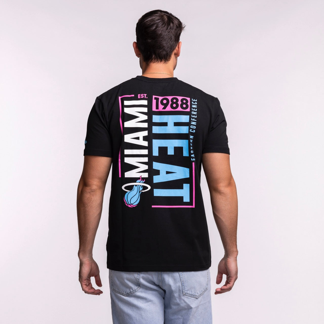 A man faces away in a New Era Miami HEAT Original Vice Nights Back Print Tee, a standard fit black cotton-spandex shirt with bold Miami Heat text, Est. 1988, Eastern Conference in pink, blue, and white plus the team logo.