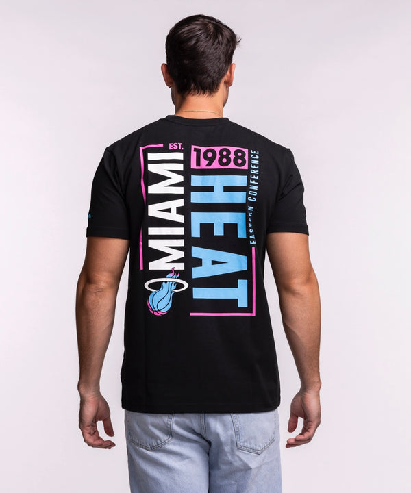 A man models the New Era Miami HEAT Original Vice Nights Back Print Tee, featuring a colorful basketball logo. He pairs it with light blue jeans and stands against a plain light background, smiling and looking to his left.