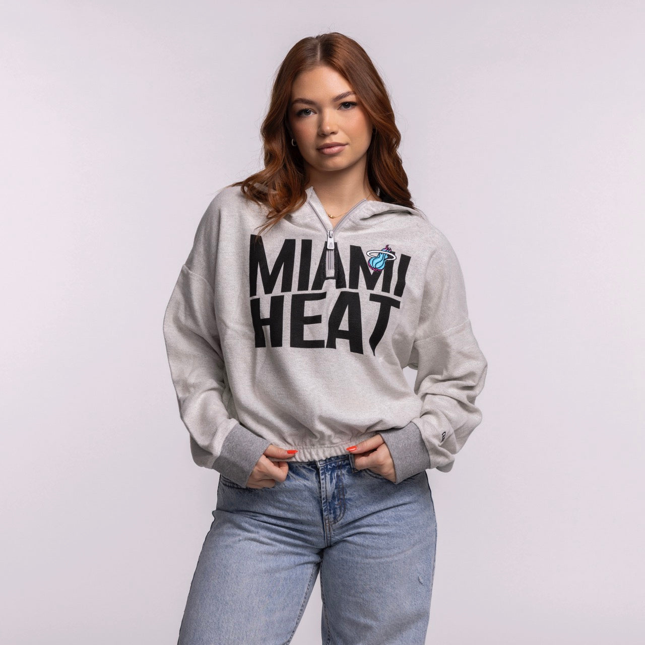 A woman with long auburn hair stands against a light background, wearing a New Era Miami HEAT Original Vice Nights 1/4 Zip Women's Hoodie in light gray and blue jeans, her hands in her pockets. Machine wash cold for care.