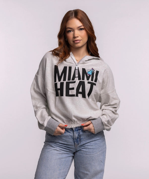 A woman with long auburn hair stands against a light background, wearing a New Era Miami HEAT Original Vice Nights 1/4 Zip Women's Hoodie in light gray and blue jeans, her hands in her pockets. Machine wash cold for care.