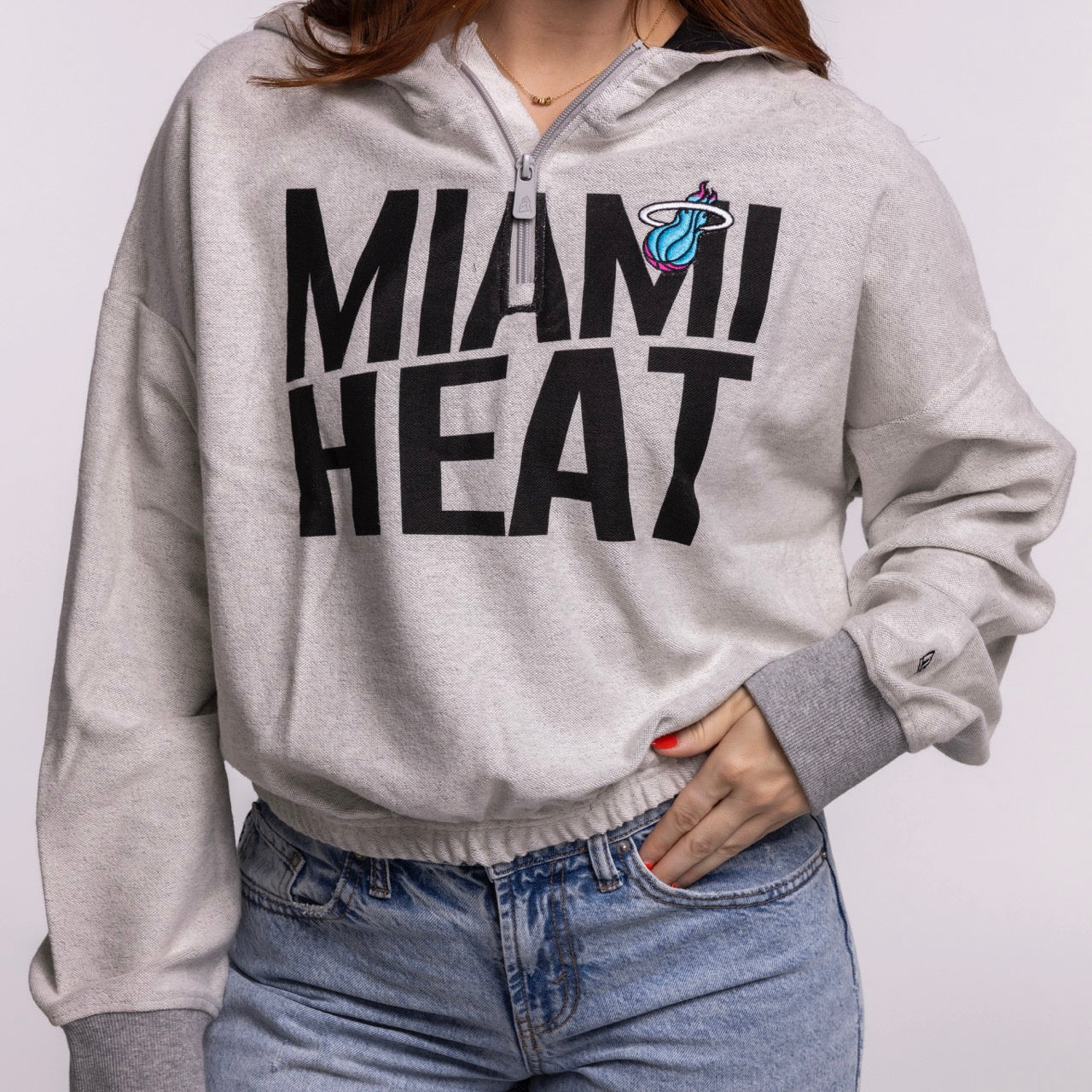 A person wears the New Era Miami HEAT Original Vice Nights 1/4 Zip Women's Hoodie—a relaxed-fit, light gray hoodie with black lettering and flaming basketball logo—paired with blue jeans, one hand in their pocket and the other on their hip.