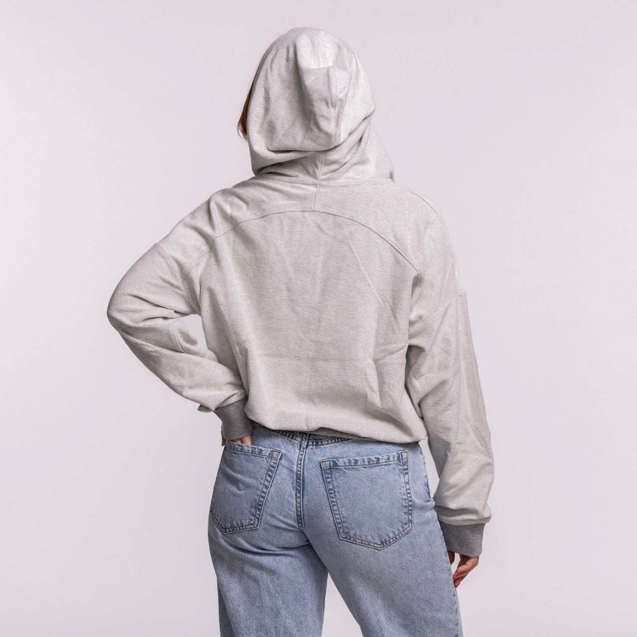 A person wearing a New Era Miami HEAT Original Vice Nights 1/4 Zip Women's Hoodie in light gray stands with their back to the camera, hood up, paired with light blue jeans against a plain light background.