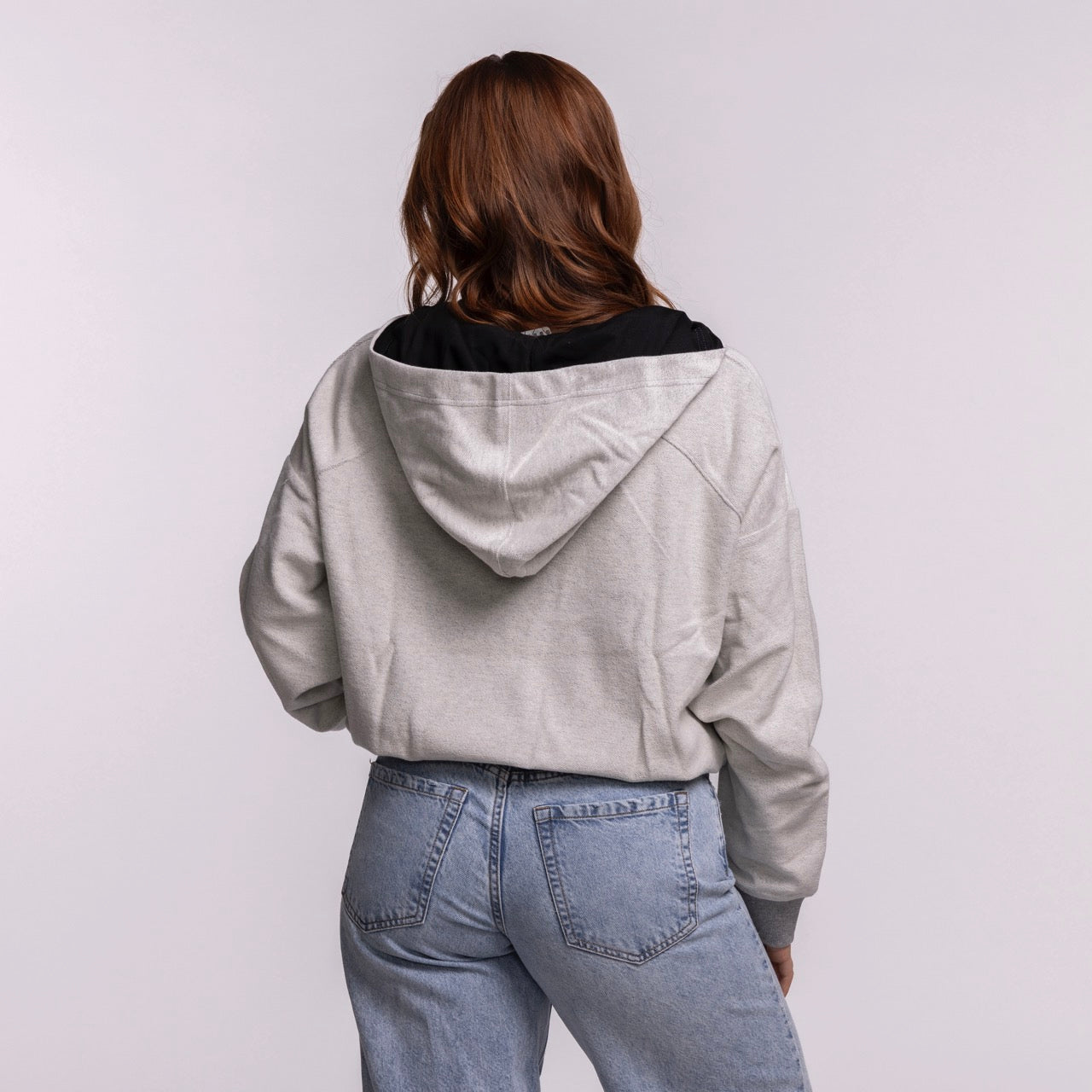 A woman with long brown hair wears the New Era Miami HEAT Original Vice Nights 1/4 Zip Women's Hoodie, featuring a relaxed cropped fit, standing in blue jeans against a plain light background. The cotton-polyester blend offers comfort and durability.