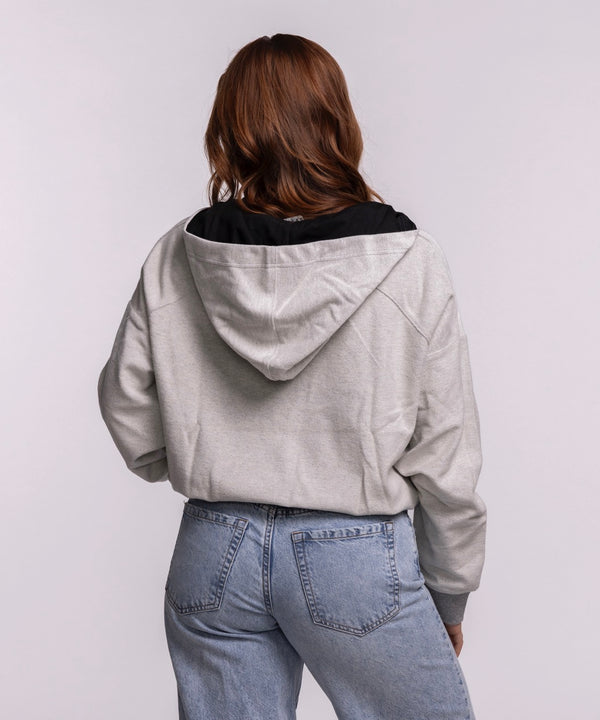 A woman with long auburn hair stands against a light background, wearing a New Era Miami HEAT Original Vice Nights 1/4 Zip Women's Hoodie in light gray and blue jeans, her hands in her pockets. Machine wash cold for care.