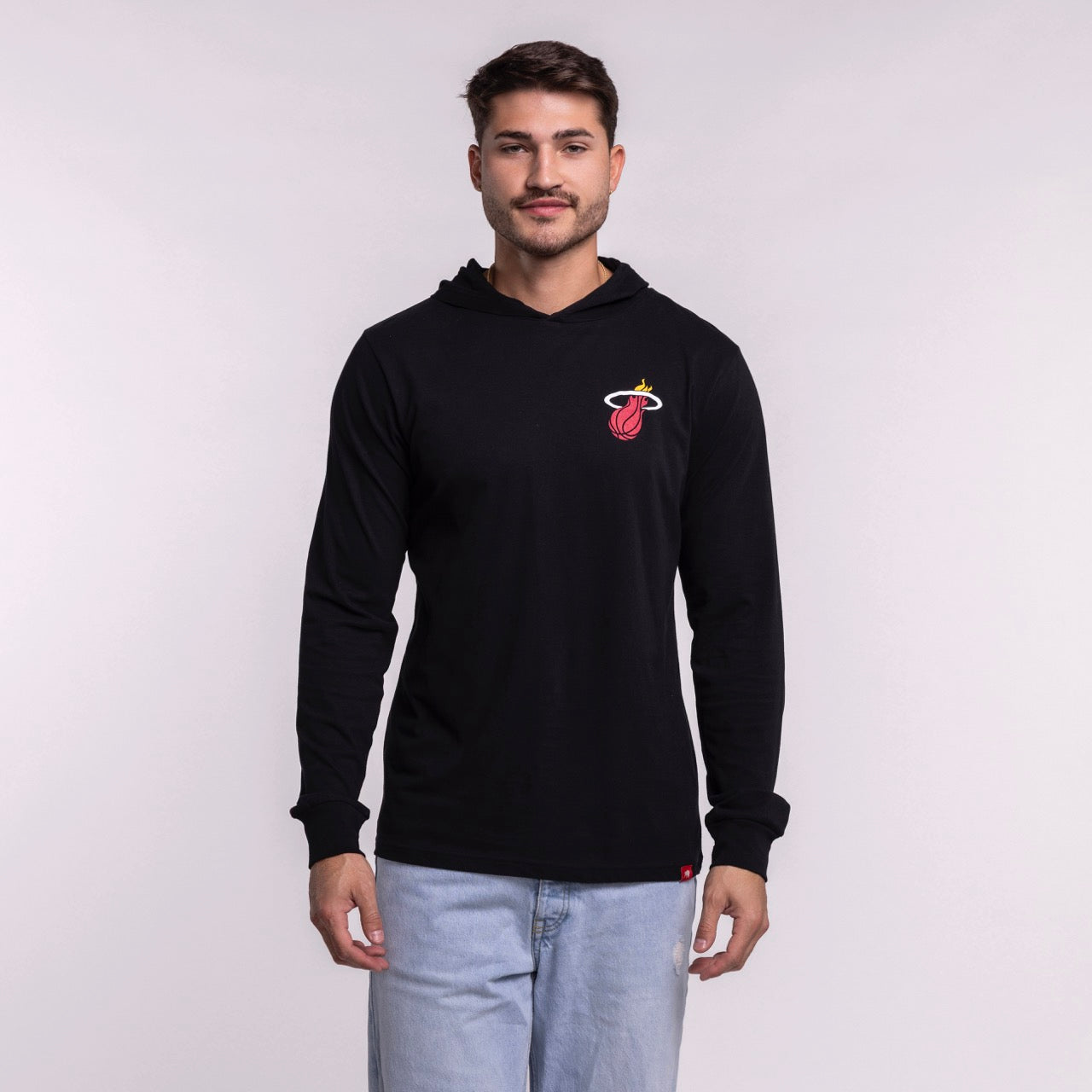 A man with short brown hair and light facial hair wears a black Sportiqe Miami HEAT Solano Logo Hoodie (100% cotton, machine washable) and light blue jeans, standing against a plain, light-colored background.