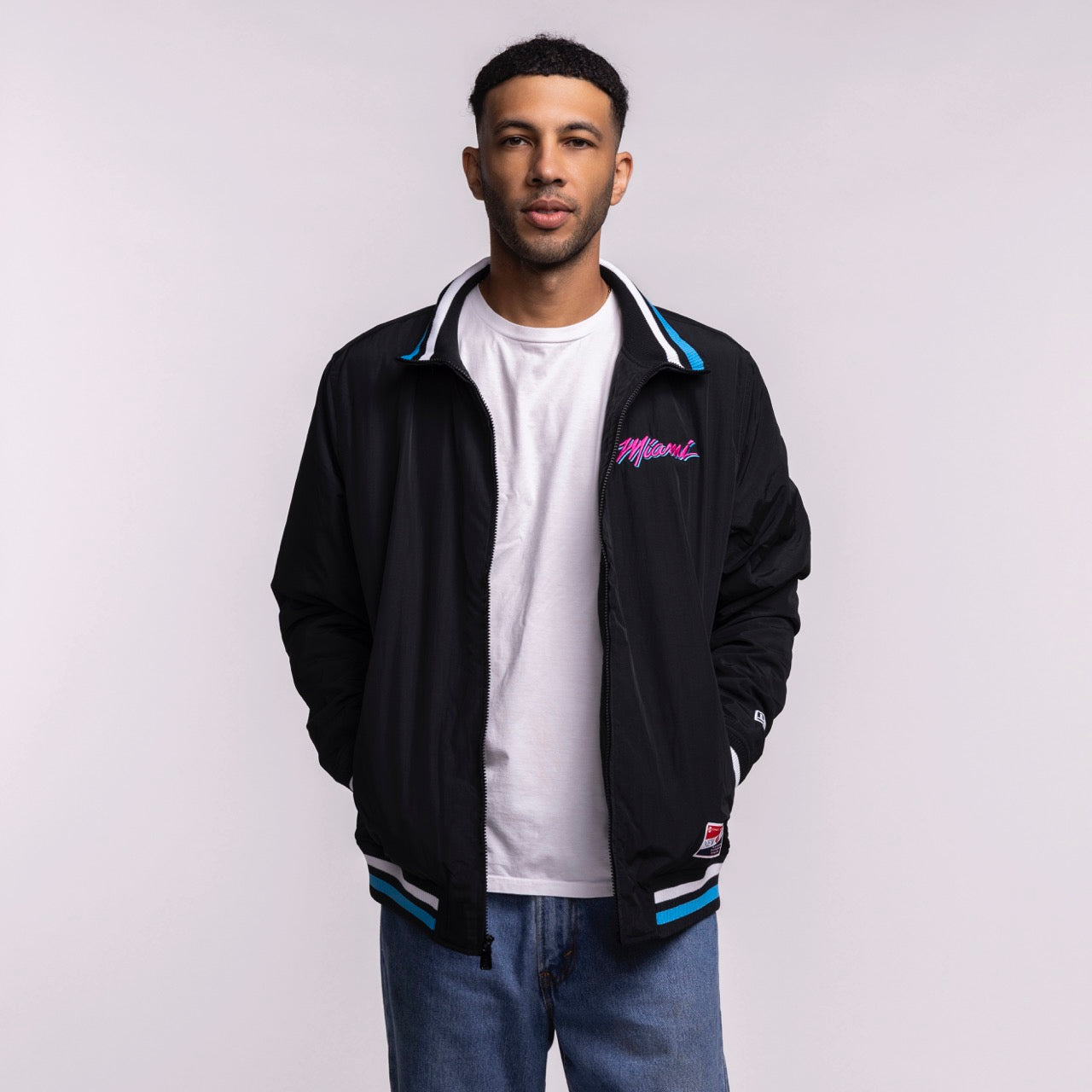 A man with short hair and a trimmed beard stands against a plain light background, wearing the New Era Miami HEAT Original Vice Nights Zip-Up Jacket over a white t-shirt and blue jeans. Hands in pockets. Standard fit; machine wash cold.