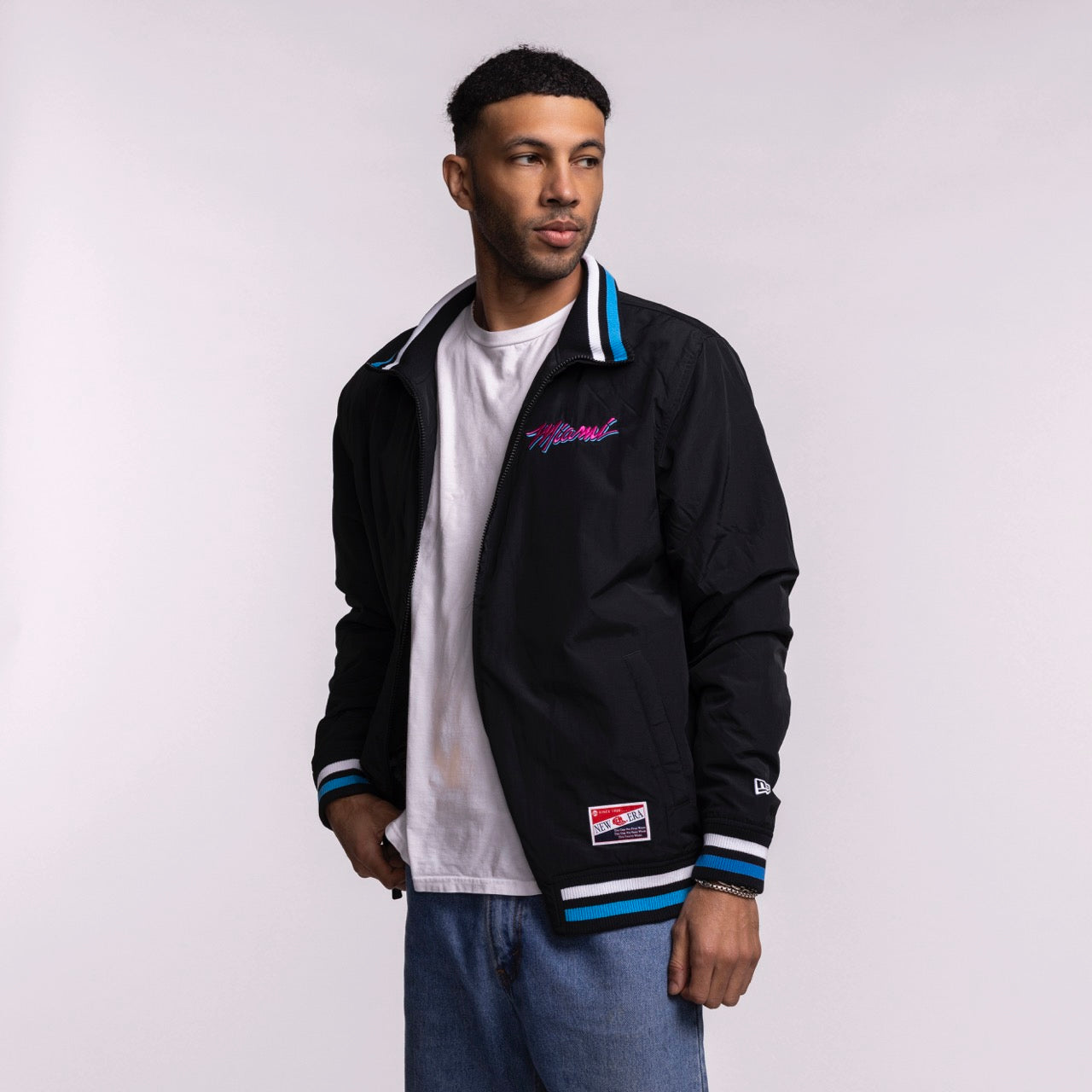 A man wears the New Era Miami HEAT Original Vice Nights Zip-Up Jacket by New Era, featuring blue and white striped cuffs and collar, over a white T-shirt and blue jeans. He stands against a plain background, looking slightly left. Machine wash cold.