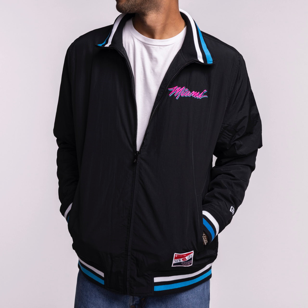 A man wearing the New Era Miami HEAT Original Vice Nights Zip-Up Jacket—a black nylon jacket with blue/white striped cuffs and collar, pink 