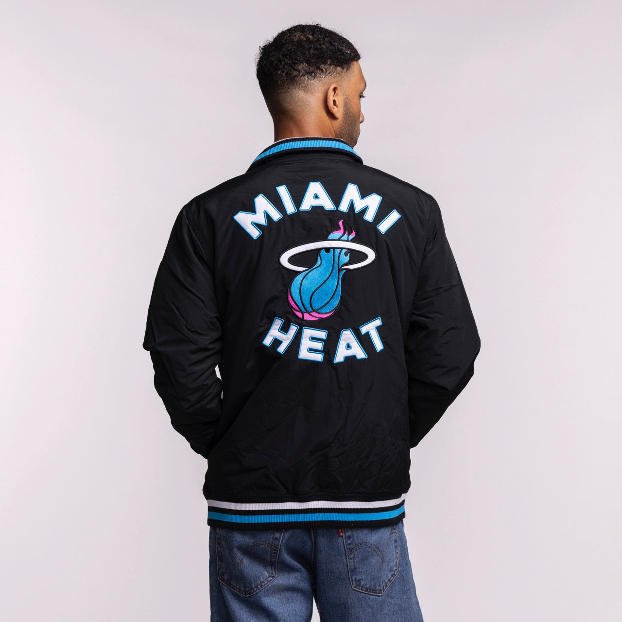A person faces away, wearing the New Era Miami HEAT Original Vice Nights Zip-Up Jacket featuring a large blue and white team logo on the back. This standard fit jacket by New Era pairs well with jeans and is machine washable for easy care.
