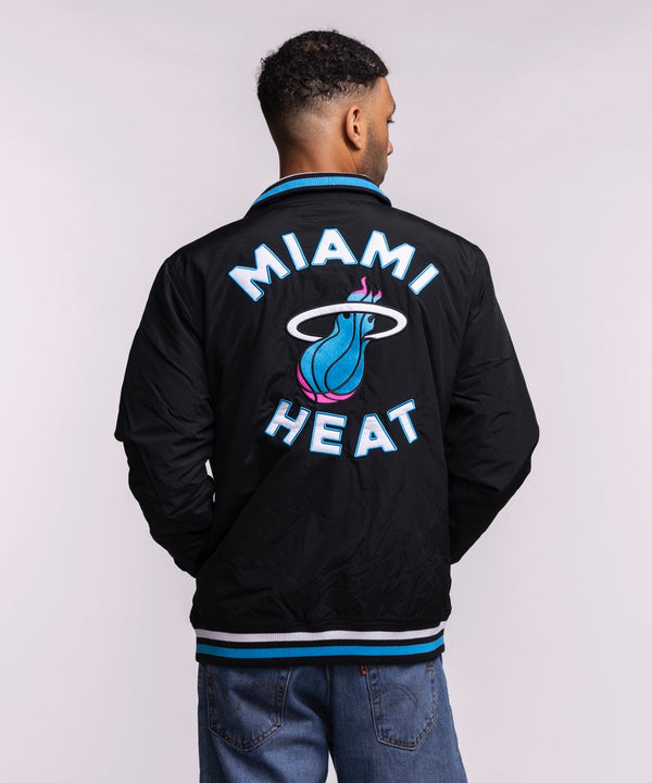 A man with short hair and a trimmed beard stands against a plain light background, wearing the New Era Miami HEAT Original Vice Nights Zip-Up Jacket over a white t-shirt and blue jeans. Hands in pockets. Standard fit; machine wash cold.