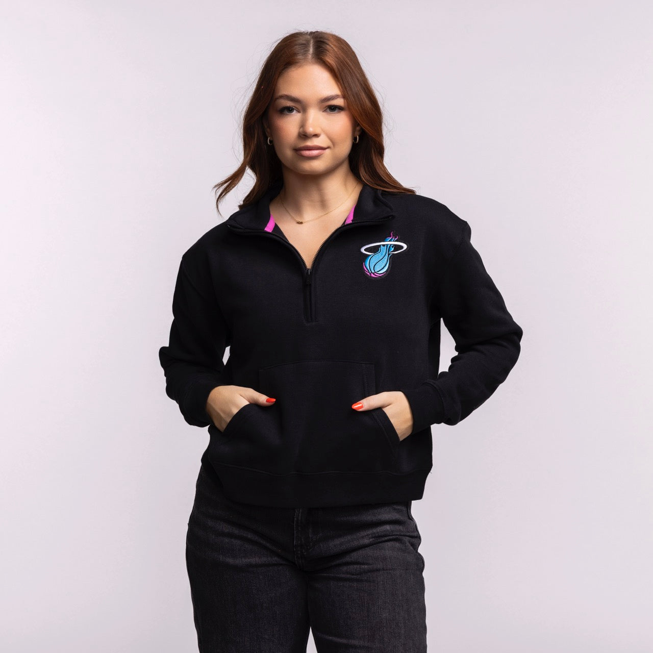 A woman with long brown hair wears the Stadium Essentials Miami HEAT Original Vice Nights 1/2 Zip Women's Jacket by Stadium Essentials, featuring a colorful Miami Heat logo, paired with dark jeans against a plain light background.