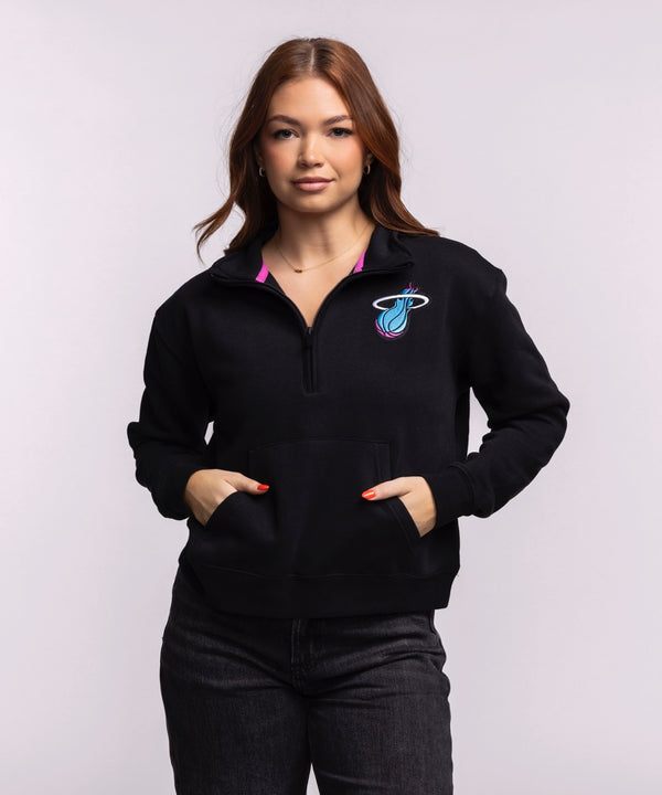 A woman with long brown hair wears the Stadium Essentials Miami HEAT Original Vice Nights 1/2 Zip Women's Jacket by Stadium Essentials, featuring a colorful Miami Heat logo, paired with dark jeans against a plain light background.