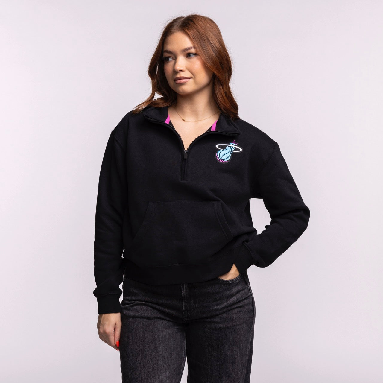A woman with long brown hair models the Stadium Essentials Miami HEAT Original Vice Nights 1/2 Zip Women's Jacket in black, paired with dark jeans. She stands against a plain light background, looking to the side with one hand in her pocket.