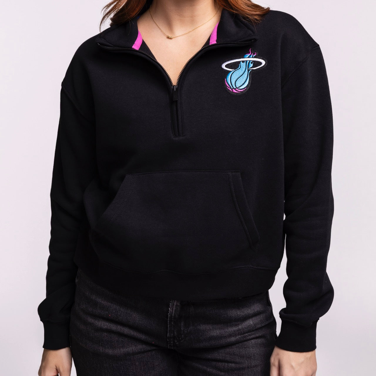 A woman wears the Stadium Essentials Miami HEAT Original Vice Nights 1/2 Zip Jacket in black with a relaxed fit and team logo, paired with dark jeans. The cotton-polyester blend offers comfort. Background is light and plain.