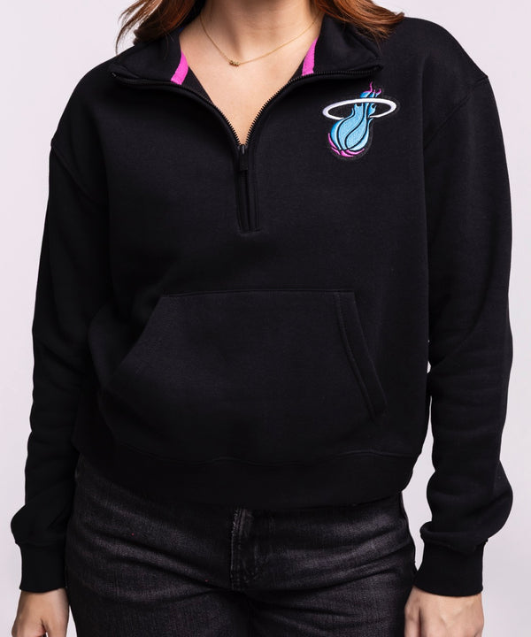 A woman with long brown hair wears the Stadium Essentials Miami HEAT Original Vice Nights 1/2 Zip Women's Jacket by Stadium Essentials, featuring a colorful Miami Heat logo, paired with dark jeans against a plain light background.