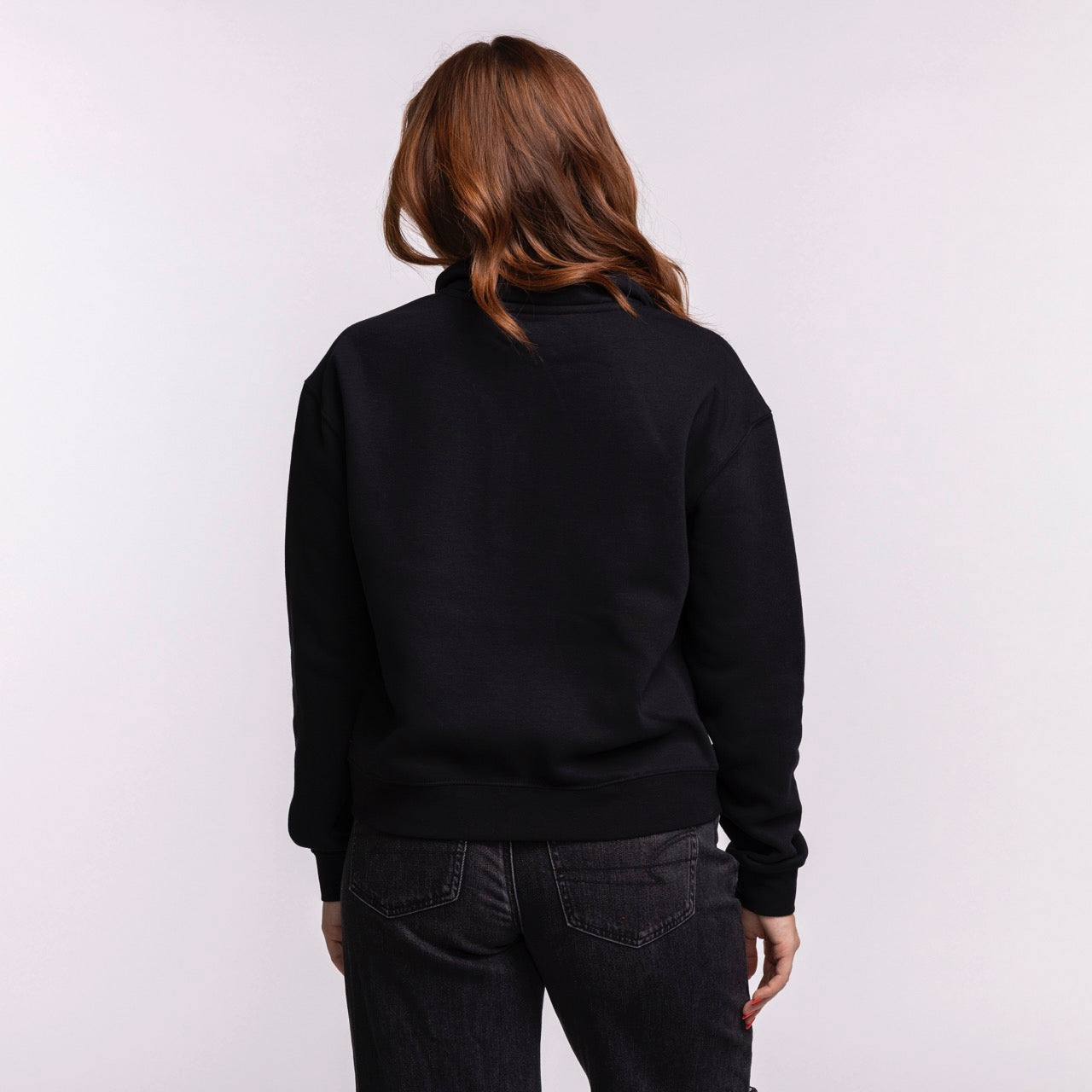 A woman with wavy, shoulder-length brown hair faces away from the camera, wearing the relaxed fit Stadium Essentials Miami HEAT Original Vice Nights 1/2 Zip Women's Jacket in black, paired with dark jeans against a light background.