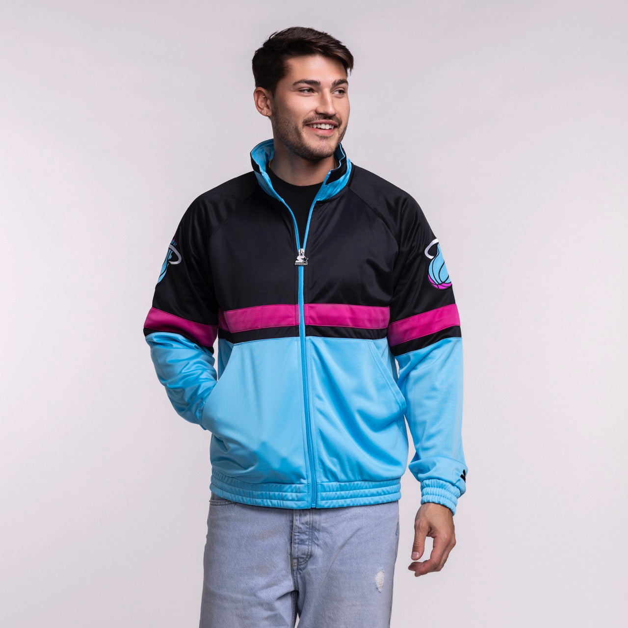 A man in a men's large G-III Sports Miami HEAT Original Vice Nights Track Jacket, featuring a sports logo on the sleeve, pairs it with light blue jeans and smiles against a plain light gray background.