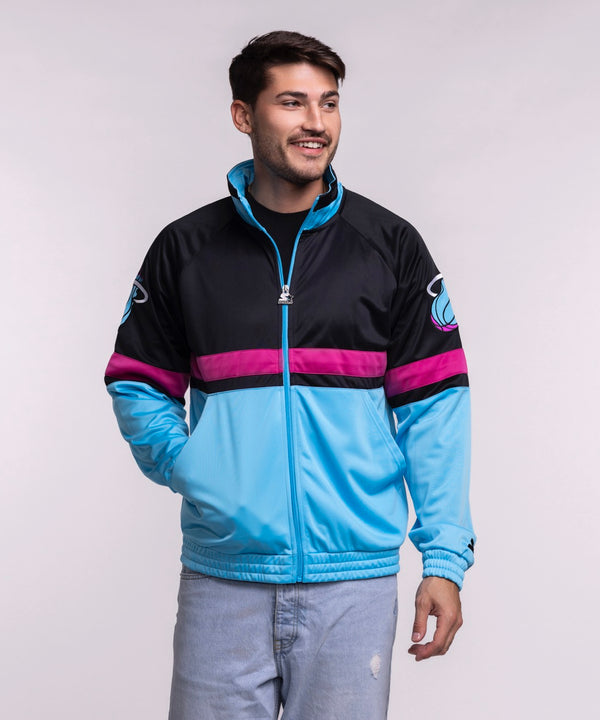 A man in a men's large G-III Sports Miami HEAT Original Vice Nights Track Jacket, featuring a sports logo on the sleeve, pairs it with light blue jeans and smiles against a plain light gray background.