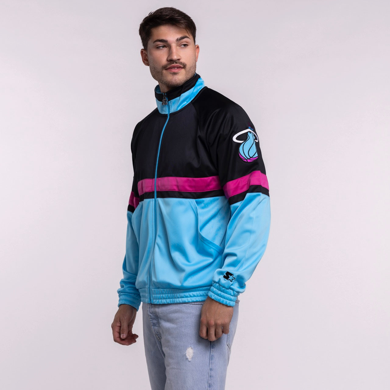 A man in a G-III Sports Miami HEAT Original Vice Nights Track Jacket—standard fit, light blue and black with a pink stripe and team logo on the sleeve—pairs it with light blue jeans, standing against a plain background, looking left.