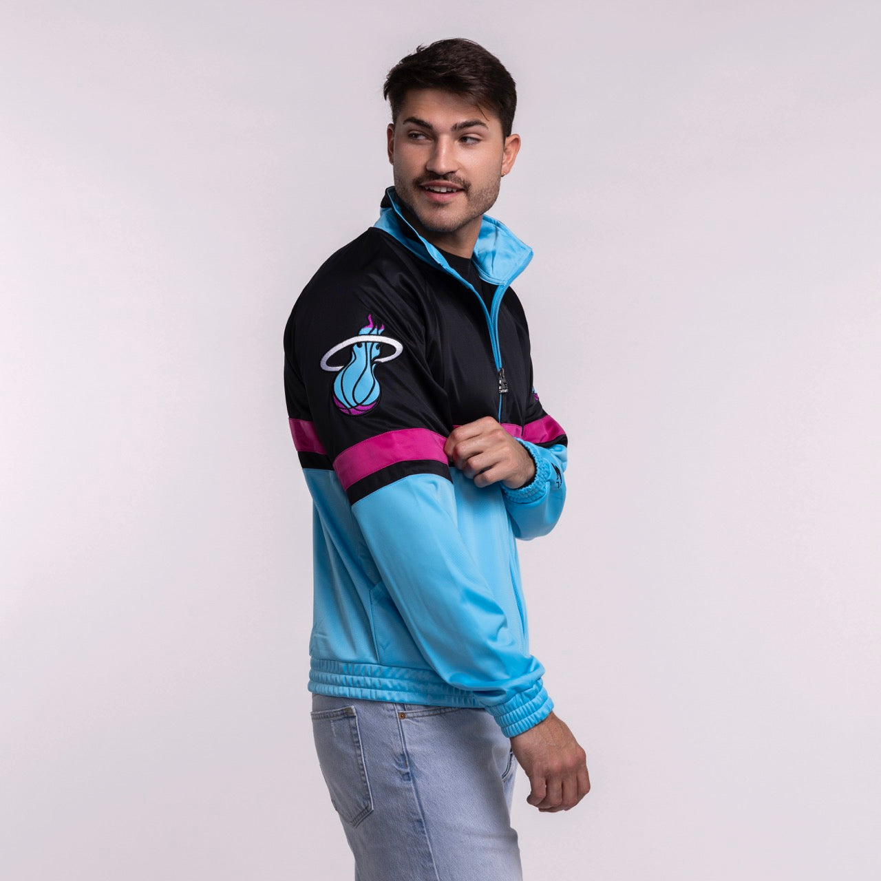 A man in light blue jeans wears the G-III Sports Miami HEAT Original Vice Nights Track Jacket by G-III Sports, featuring black with blue and magenta stripes, standing against a plain light background and smiling slightly to his right.