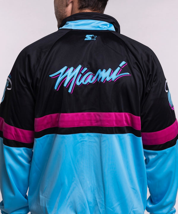 A man in a men's large G-III Sports Miami HEAT Original Vice Nights Track Jacket, featuring a sports logo on the sleeve, pairs it with light blue jeans and smiles against a plain light gray background.