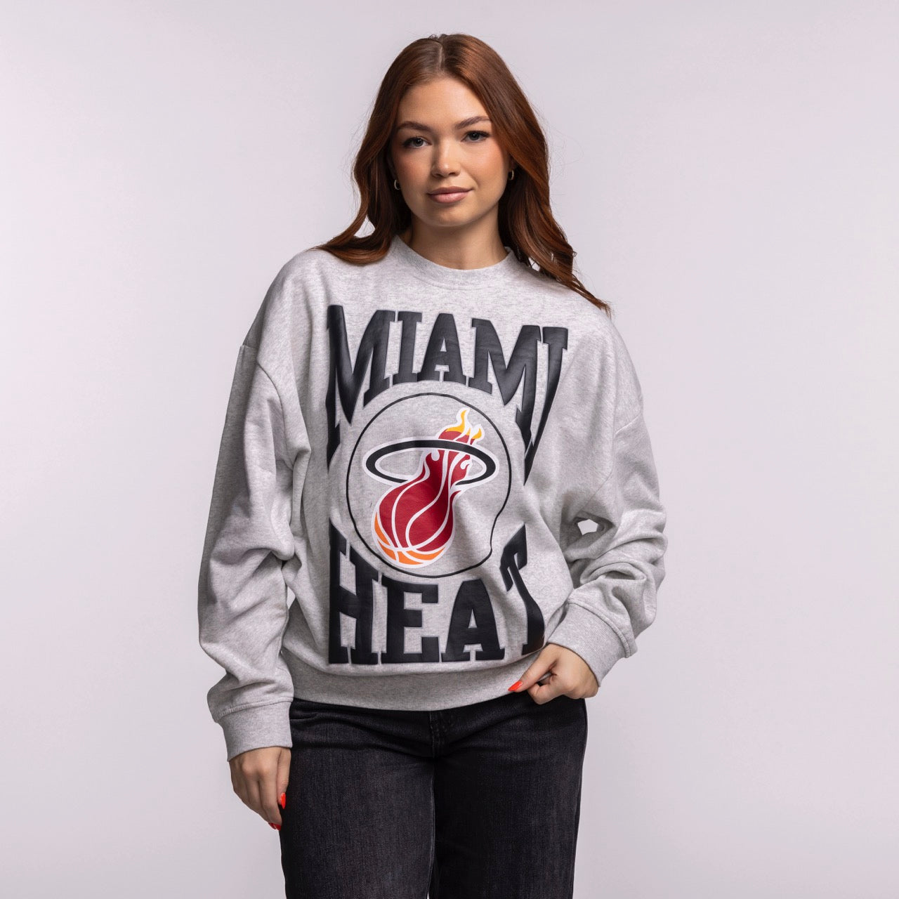 A woman with long brown hair wears the Mitchell & Ness Miami HEAT Lightweight Women's Crewneck, a relaxed fit gray sweatshirt featuring the team’s flaming logo, standing against a plain light background and looking at the camera. Machine wash cold.