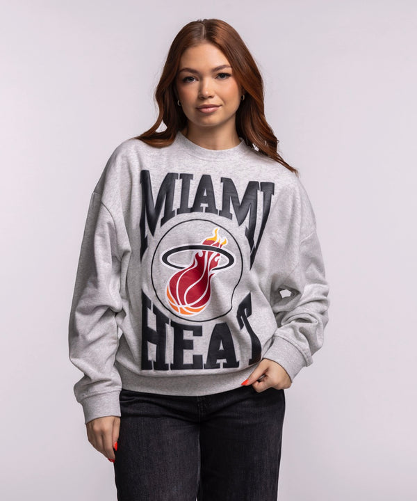 A woman with long brown hair wears the Mitchell & Ness Miami HEAT Lightweight Women's Crewneck, a relaxed fit gray sweatshirt featuring the team’s flaming logo, standing against a plain light background and looking at the camera. Machine wash cold.
