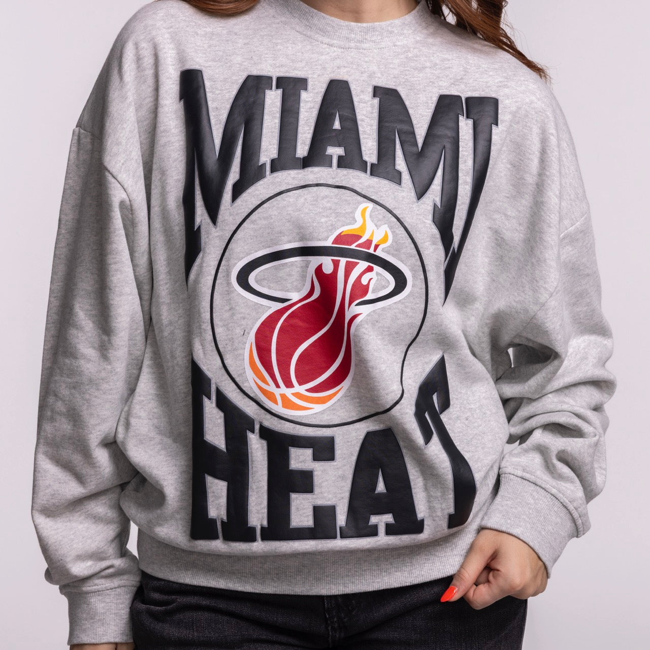 A person wears a Mitchell & Ness Miami HEAT Lightweight Women's Crewneck, a relaxed fit grey sweatshirt with a bold Miami Heat logo and name. Made of cotton polyester, it offers comfort and is machine washable. The person’s head is not fully visible.