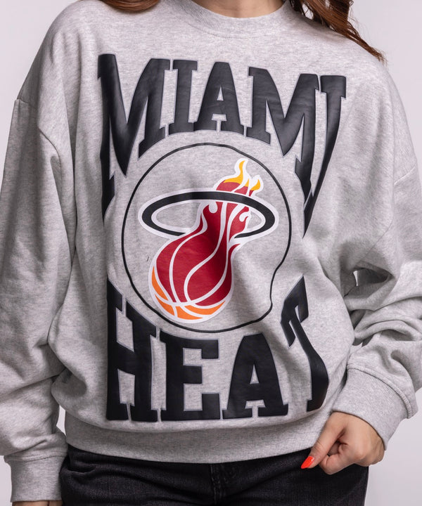 A woman with long brown hair wears the Mitchell & Ness Miami HEAT Lightweight Women's Crewneck, a relaxed fit gray sweatshirt featuring the team’s flaming logo, standing against a plain light background and looking at the camera. Machine wash cold.