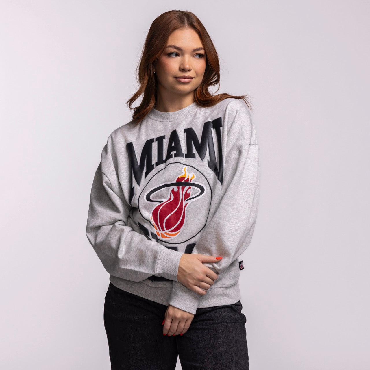 A woman with long brown hair models the Mitchell & Ness Miami HEAT Lightweight Women's Crewneck in gray, paired with dark pants. She stands against a plain, light background and gazes to the side with a relaxed expression.
