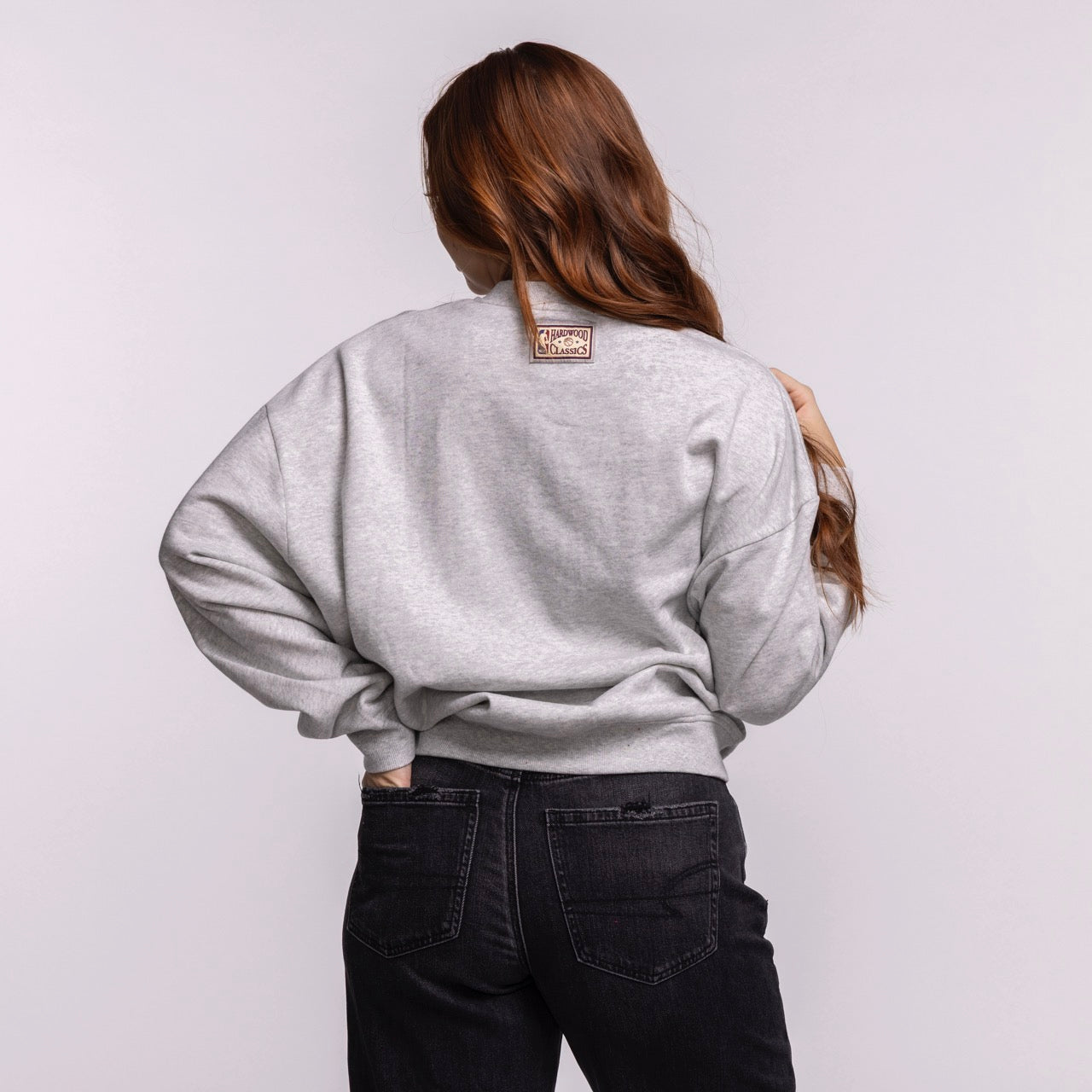 A person with long brown hair faces away, wearing a light gray Mitchell & Ness Miami HEAT Lightweight Women's Crewneck and black jeans. The relaxed fit sweatshirt has a small rectangular patch on the upper back. The background is plain and light.